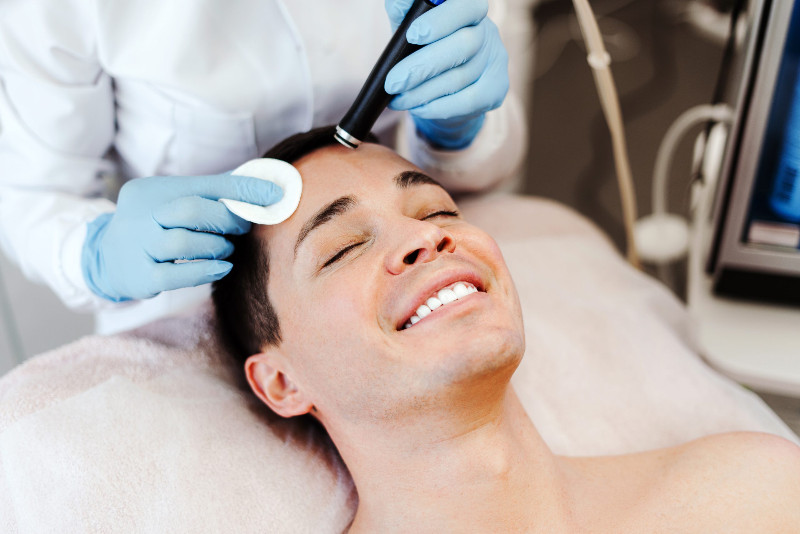 Profession cosmetologist making vacuum facial skin cleaning for smiling man in beauty salon. Handsome male lying with closed eyes. Cosmetology procedure concept